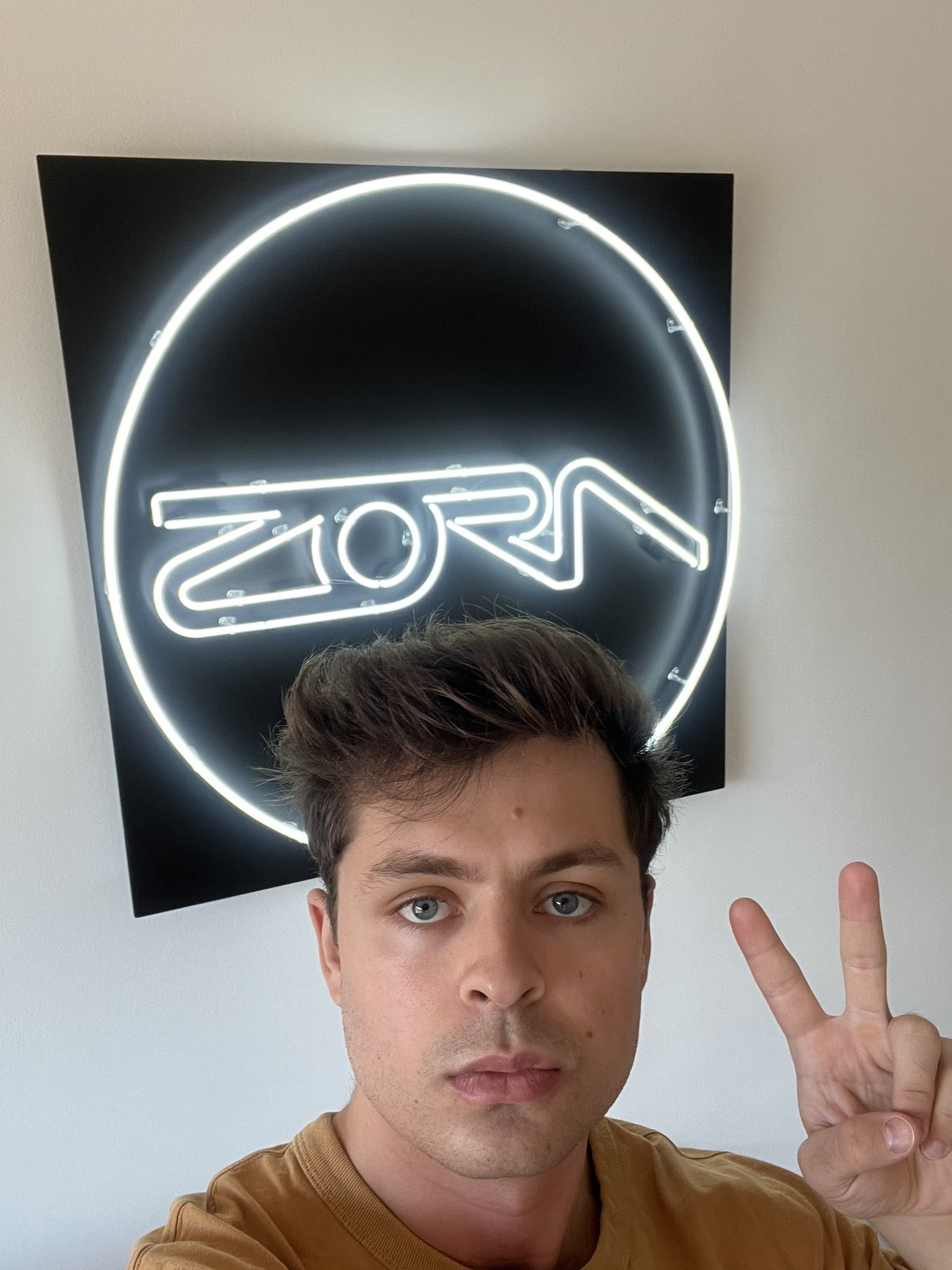 selfie of FELT Zine member Jawn Billettes. He is looking directly into the camera with a serious expression, whilst holding up a piece sign with two fingers. He is standing in front of a neon sign advertising the web3 company Zora.